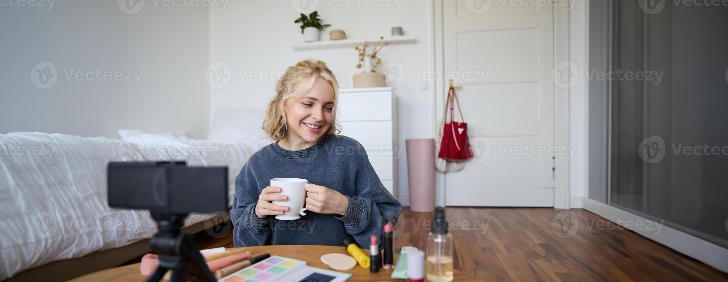 Portrait of cute blond woman, content maker, vlogger recording on digital camera in bedroom, drinking cup of tea and talking to audience photo