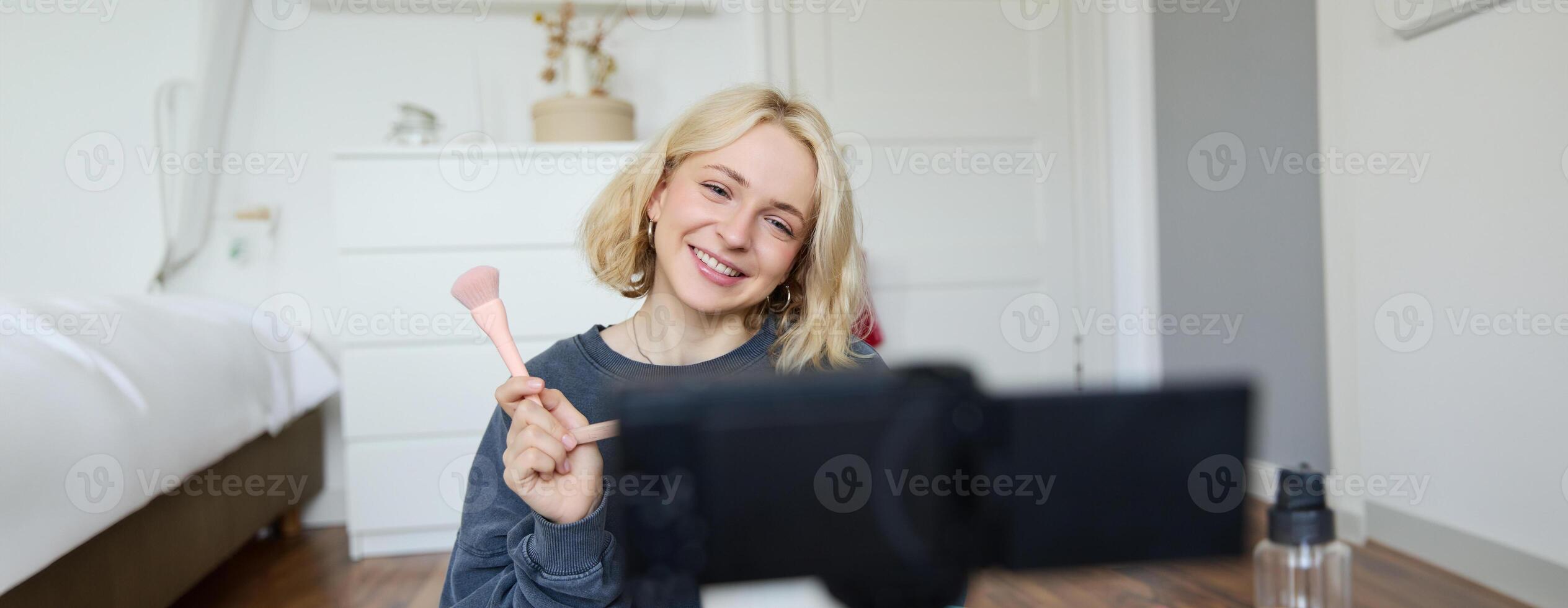 Close up portrait of happy young beauty blogger, records lifestyle vlog in her room, using camera with stabiliser, shows makeup brush and cosmetics photo