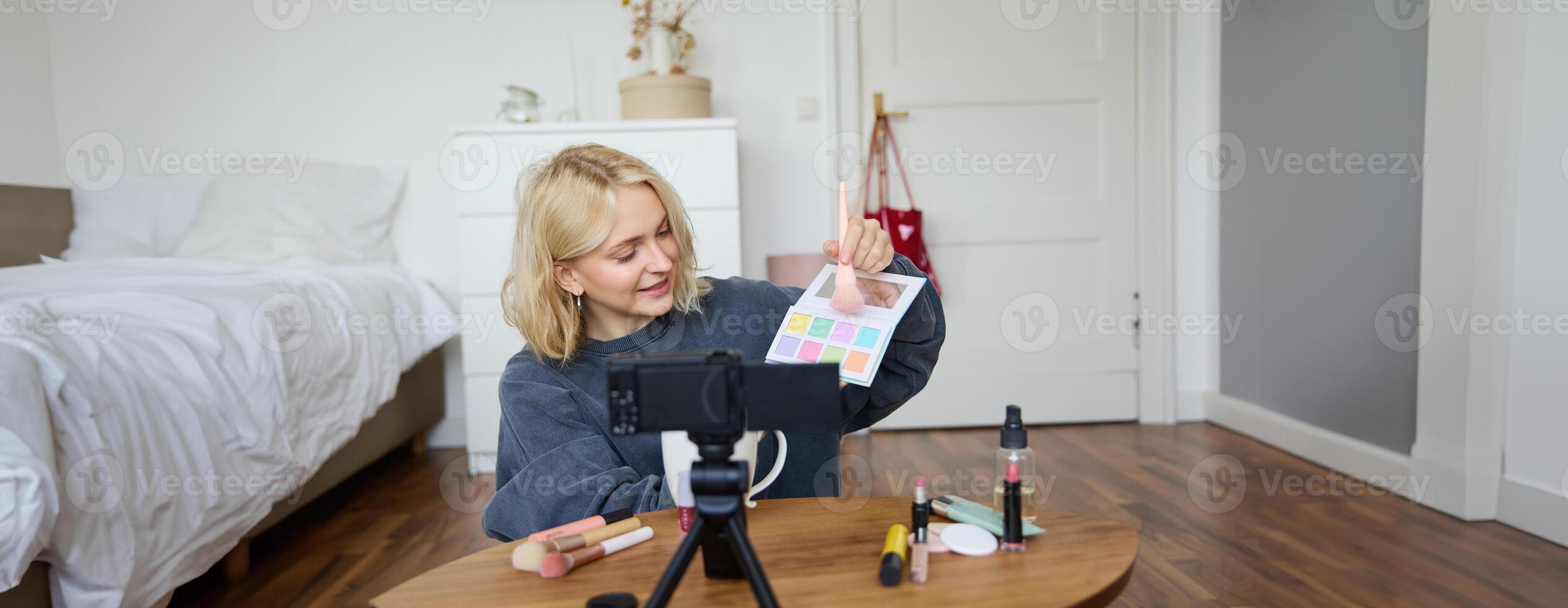 Portrait of smiling blond woman, teenage girl records a vlog on camera, vlogger shows eyeshadows, tutorial how to put makeup on, sits in her room on floor near coffee table photo
