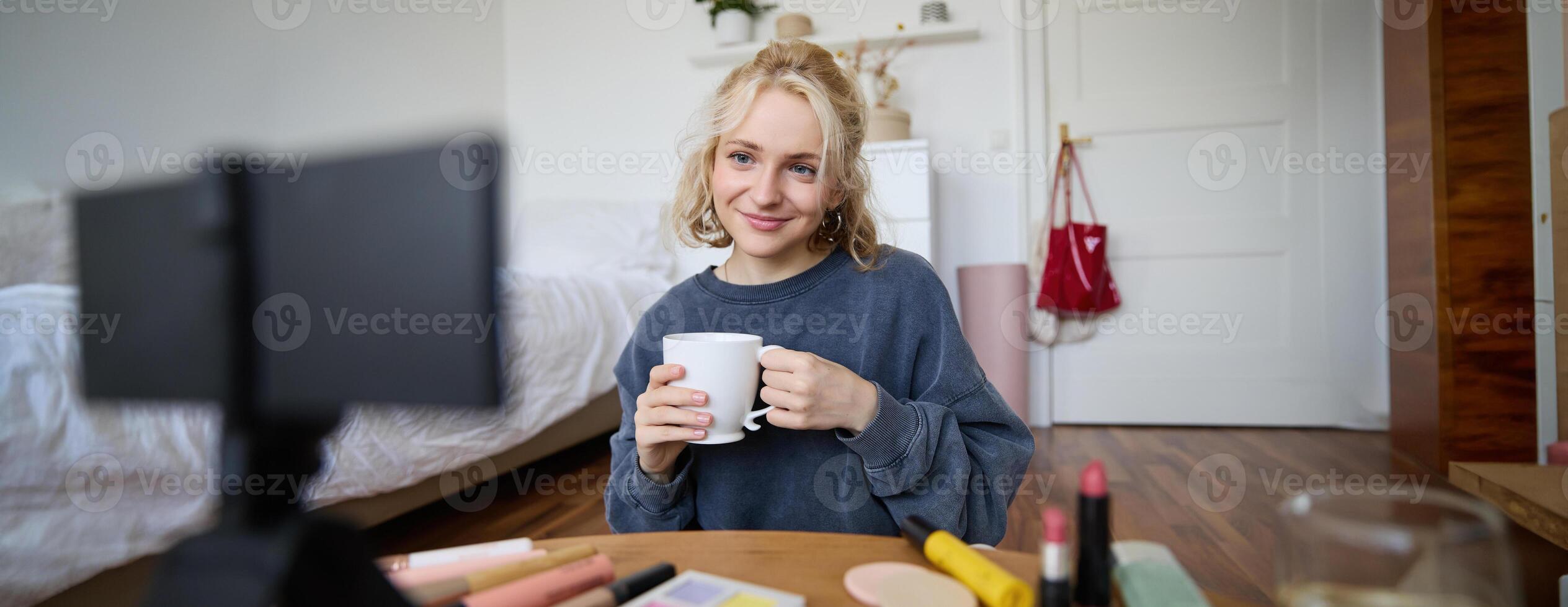 Portrait of cute blond woman, content maker, vlogger recording on digital camera in bedroom, drinking cup of tea and talking to audience photo