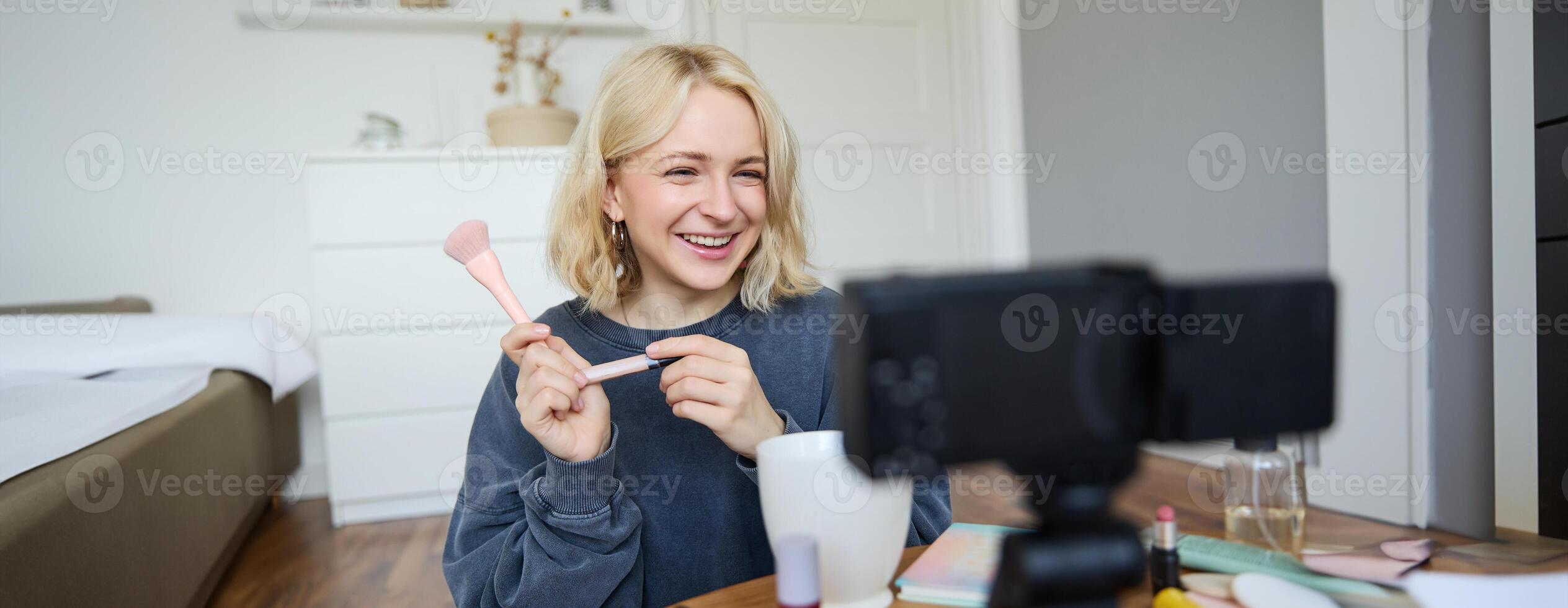 Close up portrait of happy young beauty blogger, records lifestyle vlog in her room, using camera with stabiliser, shows makeup brush and cosmetics photo