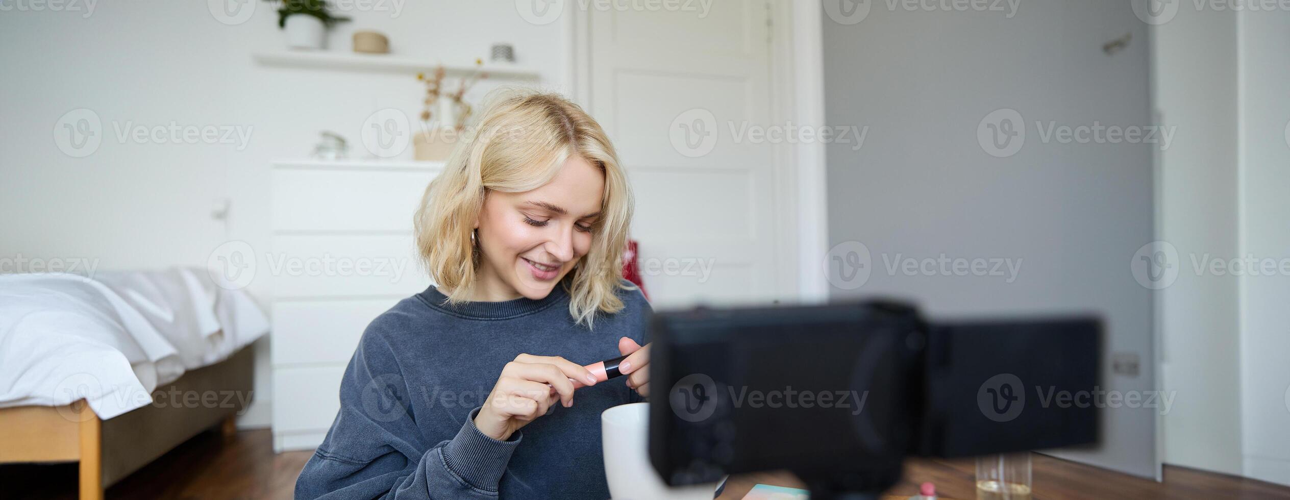 Close up portrait of smiling young woman with camera in her room, showing mascara, reviewing beauty products for social media account. Beauty blogger creates vlog about makeup photo