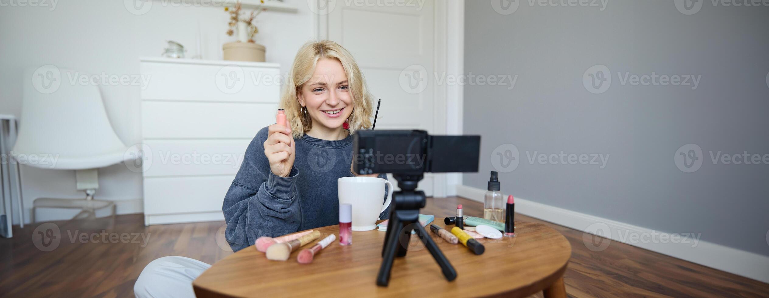 Portrait of beautiful, smiling blond woman, girl recording of her makeup tutorial for social media, vlogger sitting on floor in her room, using stabiliser to create content, reviewing mascara photo