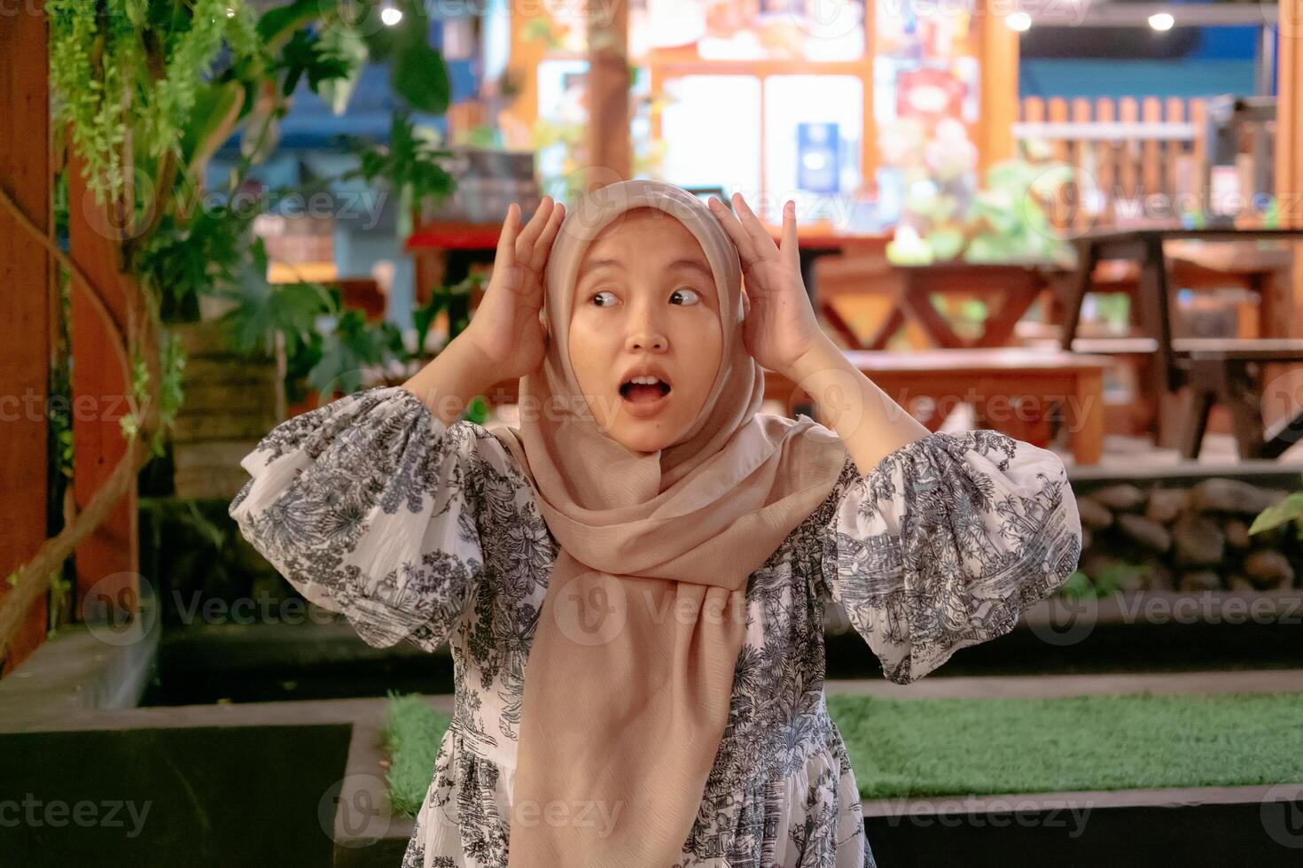 A young Asian woman wearing a hijab with a shocked expression against the backdrop of a cafe photo