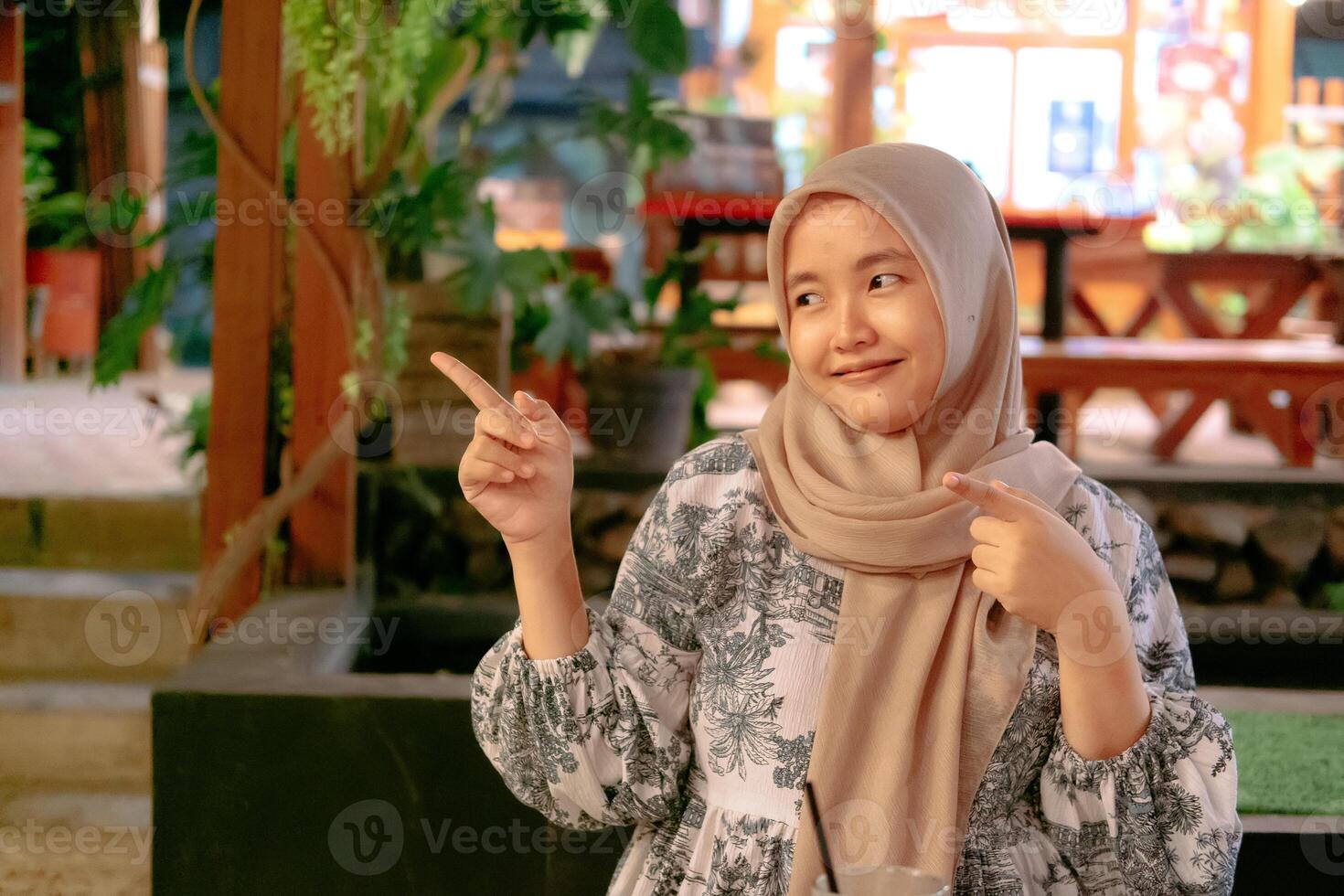 A young Asian woman wearing a hijab with a pointing expression against the background of a cafe photo