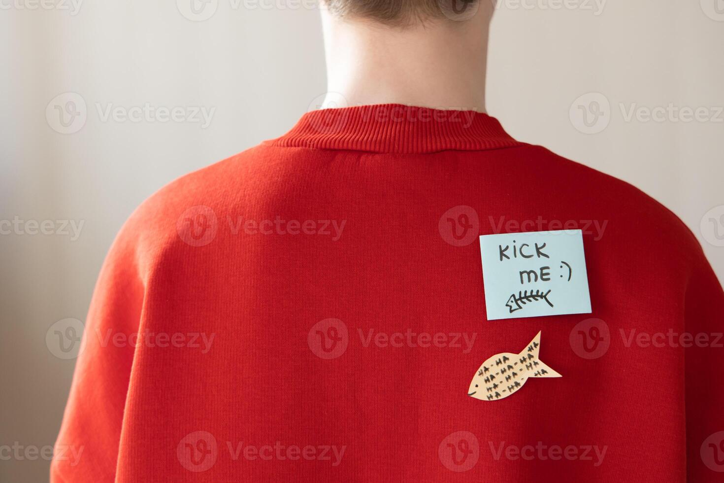 Boy in red hoodie with a paper attached with tape to his back with the handwriting text kick me. Fools day joke photo