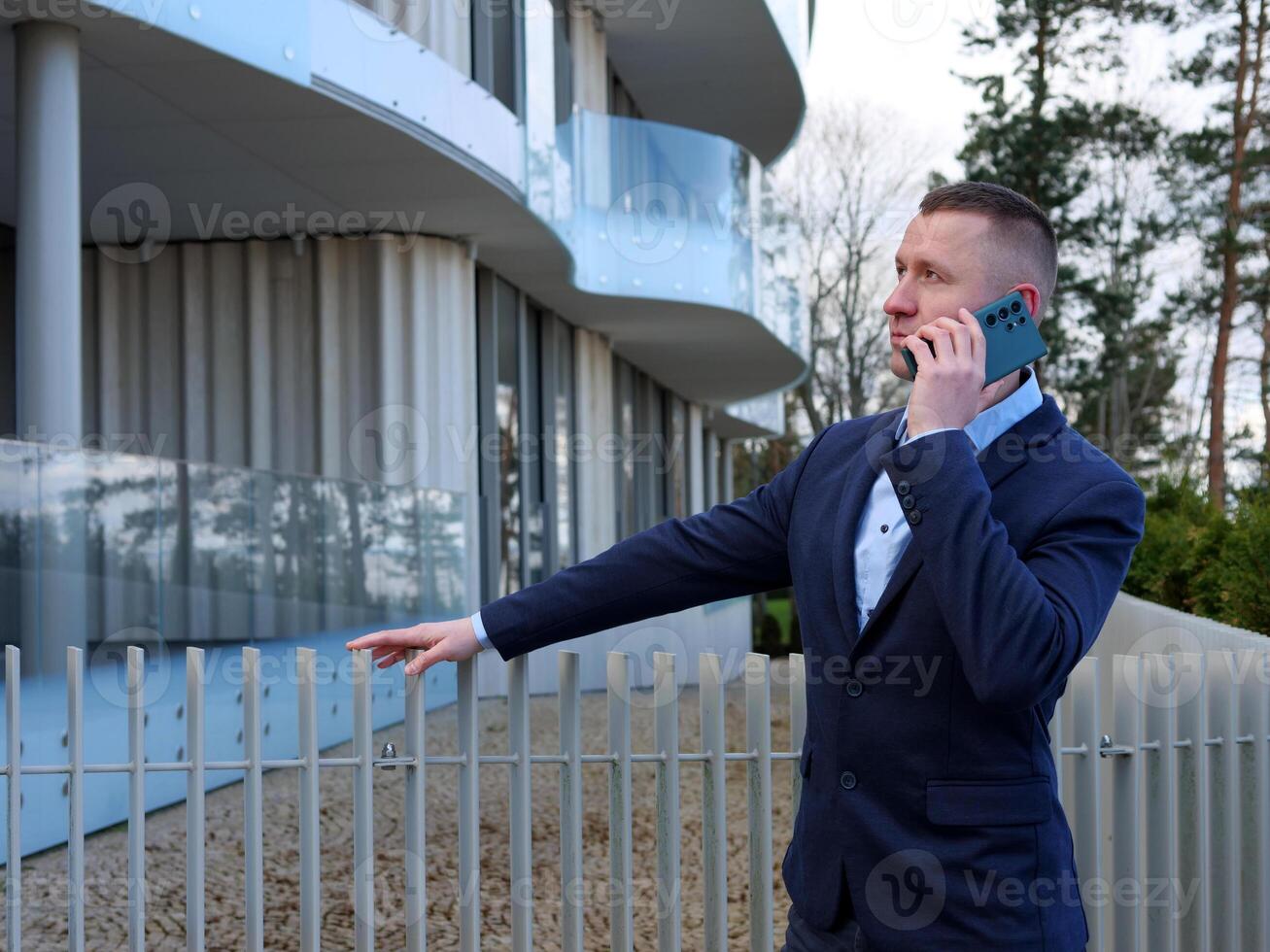 A Businessman engages in a phone conversation outside an office building, dressed in formal business attire. The Businessman's professional demeanor is evident as he discusses important matters. photo