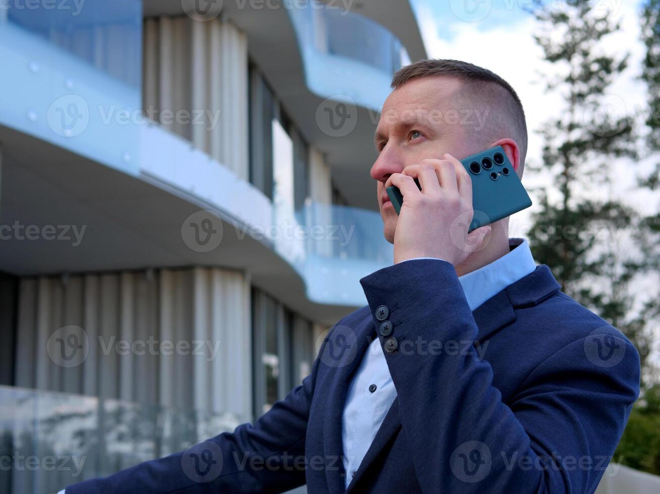 A Businessman engages in a phone conversation outside an office building, dressed in formal business attire. The Businessman's professional demeanor is evident as he discusses important matters. photo