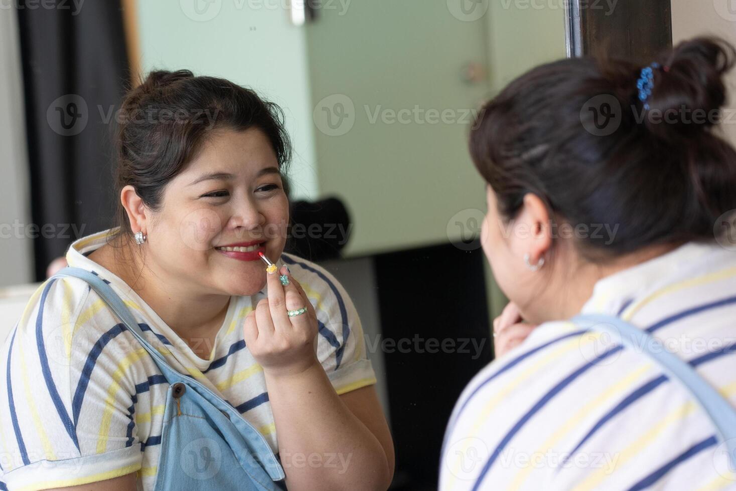 Portrait of Asian Plus Size Woman Looking at Mirror and Applying Lipstick on Lips. Concept of Postive Body Figure photo