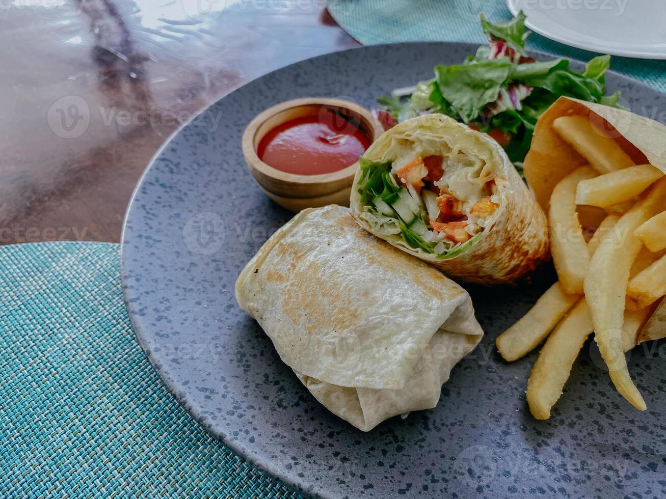 Tortilla wrap with fried chicken meat and vegetables served with sauce and french fries on a wooden table. photo