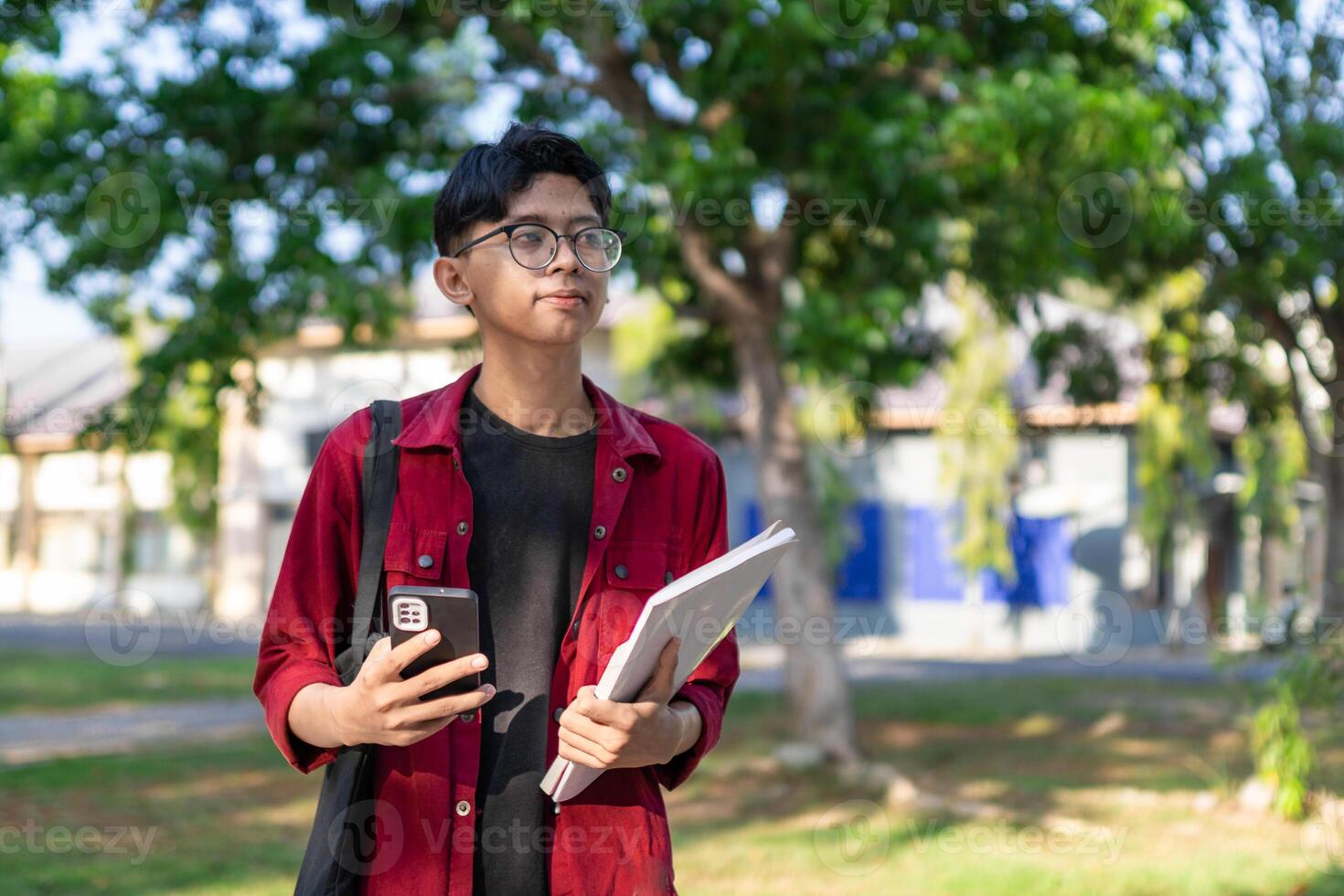 Young Asian college student using smartphone with happy expression. A male smiling while holding his phone and books at the public park. copy space photo
