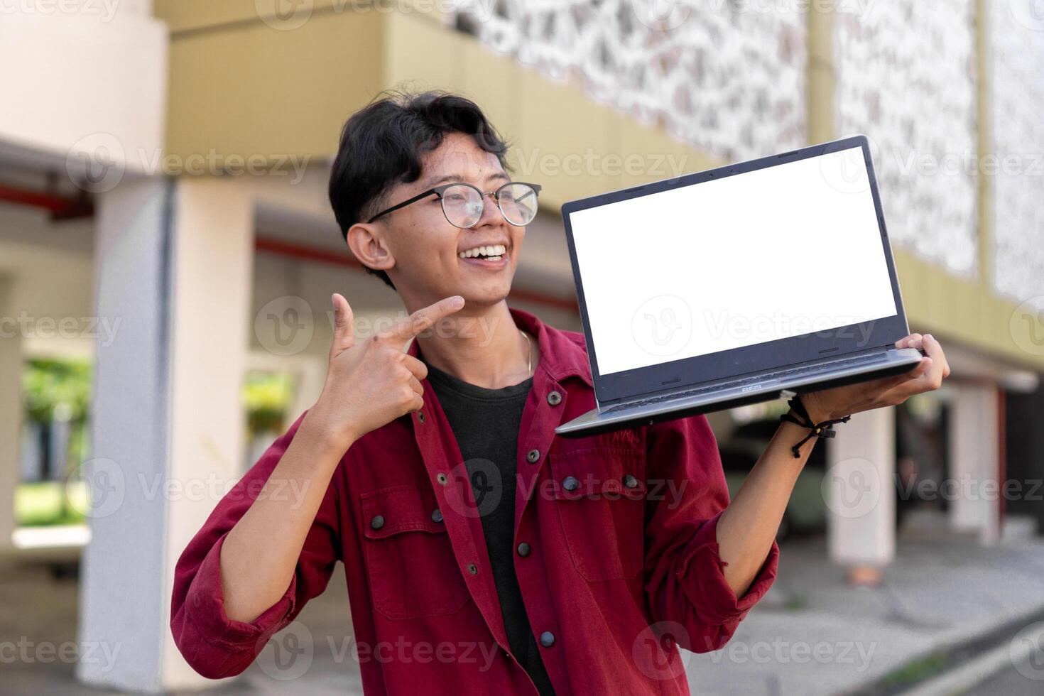 Young Asian man wearing red shirt smiling and showing white laptop screen for advertisement. Concept of people lifestyle. Happy man holding blank laptop screen with blurred background. photo