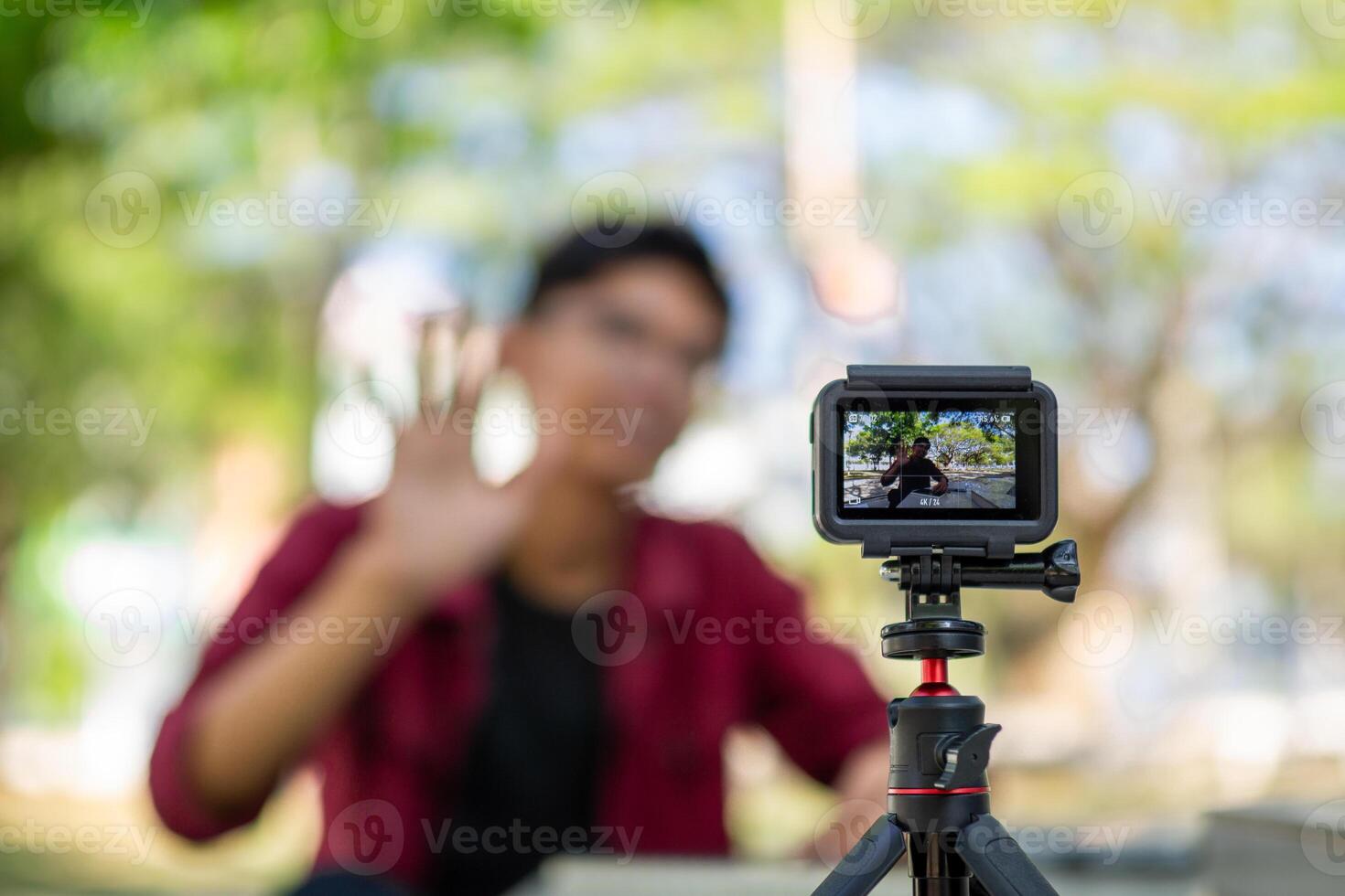 Asian man recording and talking to camera. Young man vlogging and taking selfie with action camera at public park. Action camera recording a man with blurry background photo