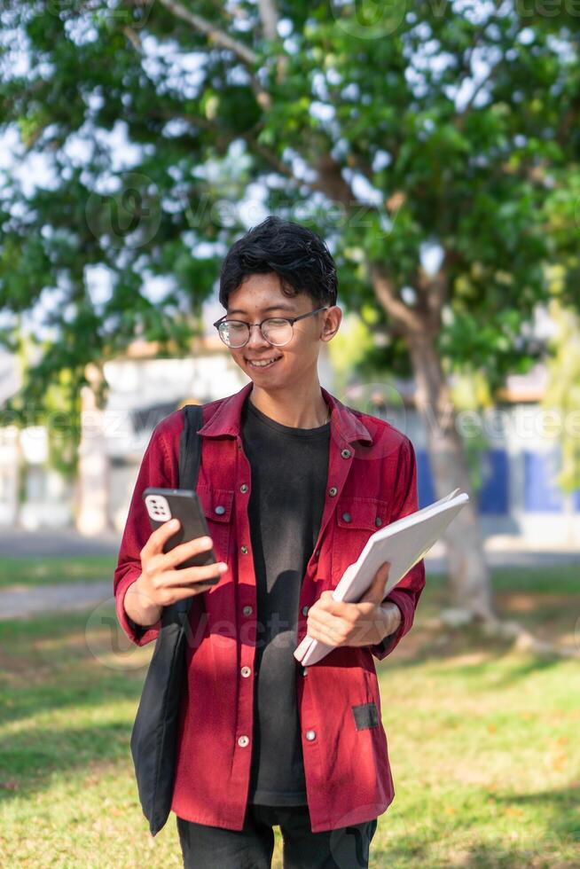 Young Asian college student using smartphone with happy expression. A male smiling while holding his phone and books at the public park. copy space photo