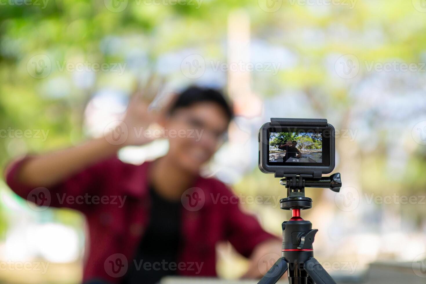 Asian man recording and talking to camera. Young man vlogging and taking selfie with action camera at public park. Action camera recording a man with blurry background photo