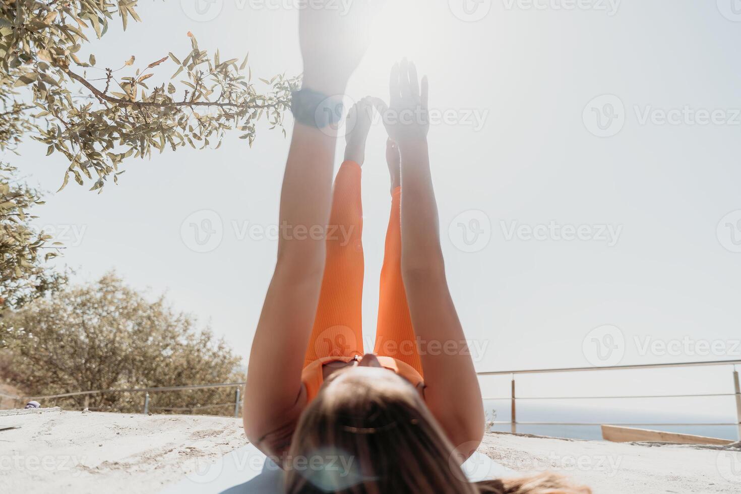 Fitness woman sea. Happy middle aged woman in orange sportswear exercises morning outdoors on yoga mat with laptop in park over ocean beach. Female fitness pilates yoga routine. Healthy lifestyle. photo