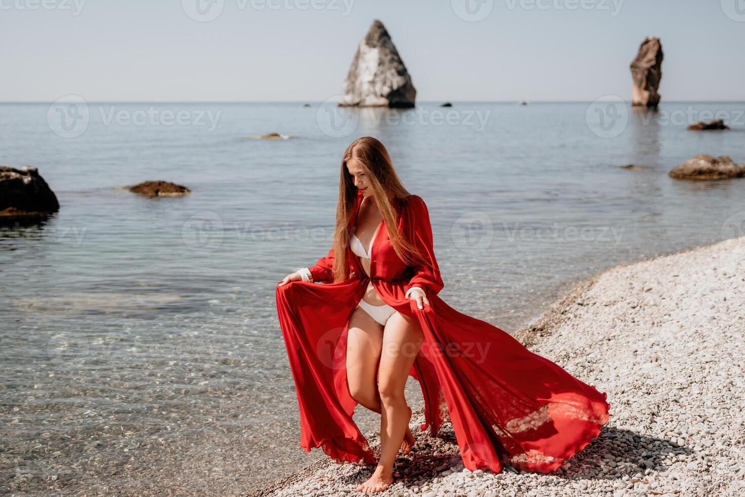 Woman travel sea. Happy tourist in red dress enjoy taking picture outdoors for memories. Woman traveler posing on the rock at sea bay surrounded by volcanic mountains, sharing travel adventure journey photo