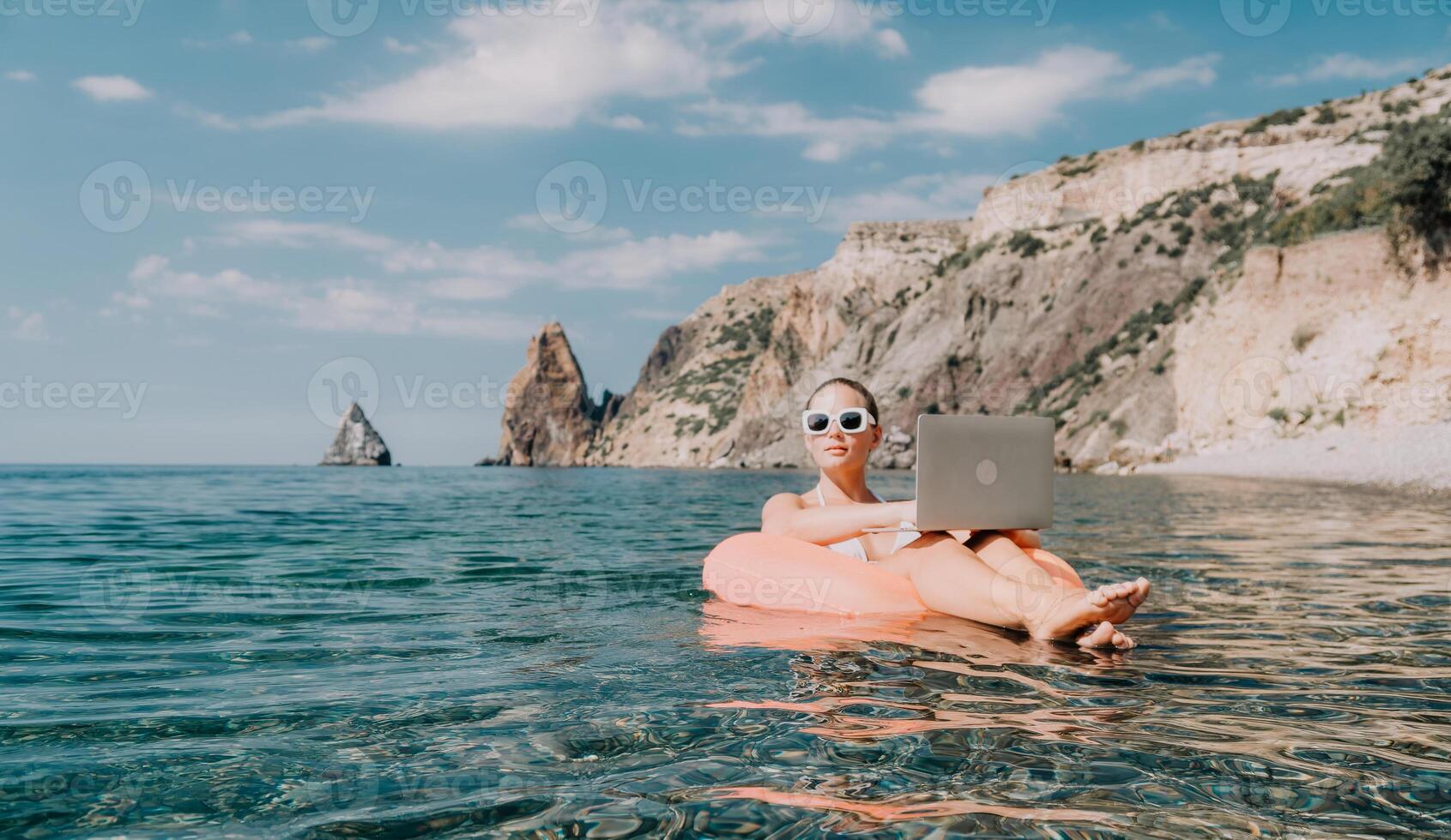 Woman freelancer works on laptop swimming in sea on pink inflatable ring. Happy tourist in sunglasses floating on inflatable donut and working on laptop computer in calm ocean. Remote working anywhere photo