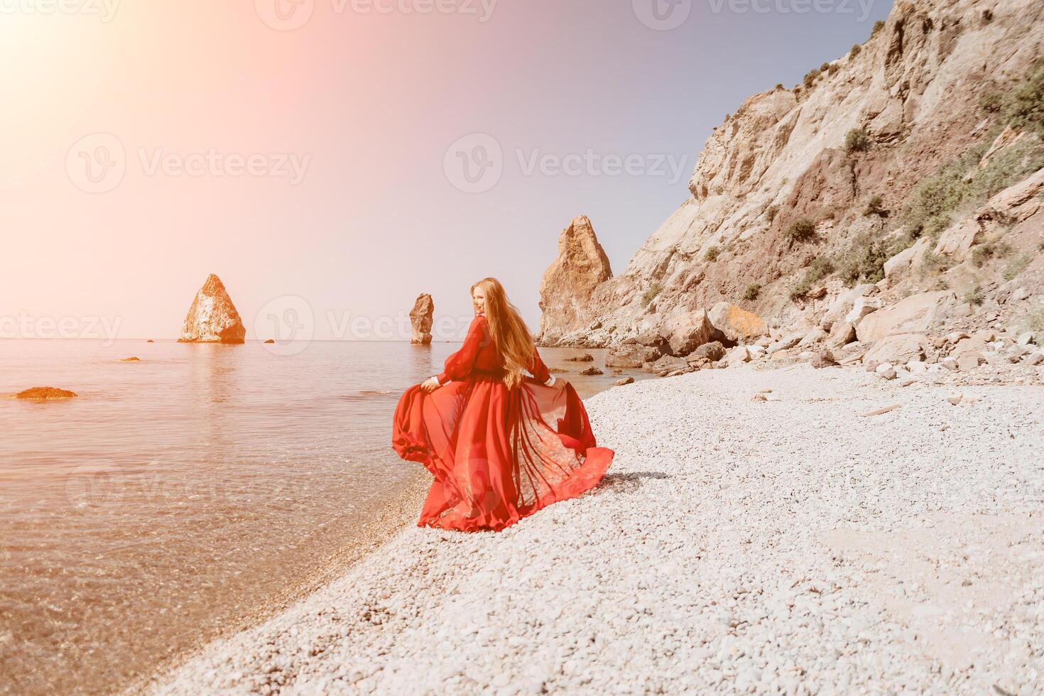Woman travel sea. Happy tourist in red dress enjoy taking picture outdoors for memories. Woman traveler posing on the rock at sea bay surrounded by volcanic mountains, sharing travel adventure journey photo