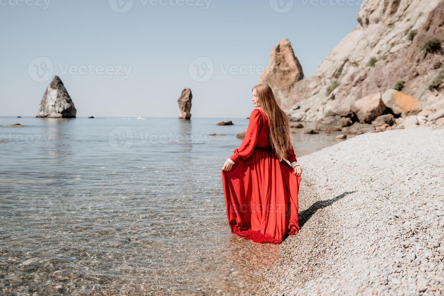 Woman travel sea. Happy tourist in red dress enjoy taking picture outdoors for memories. Woman traveler posing on the rock at sea bay surrounded by volcanic mountains, sharing travel adventure journey photo