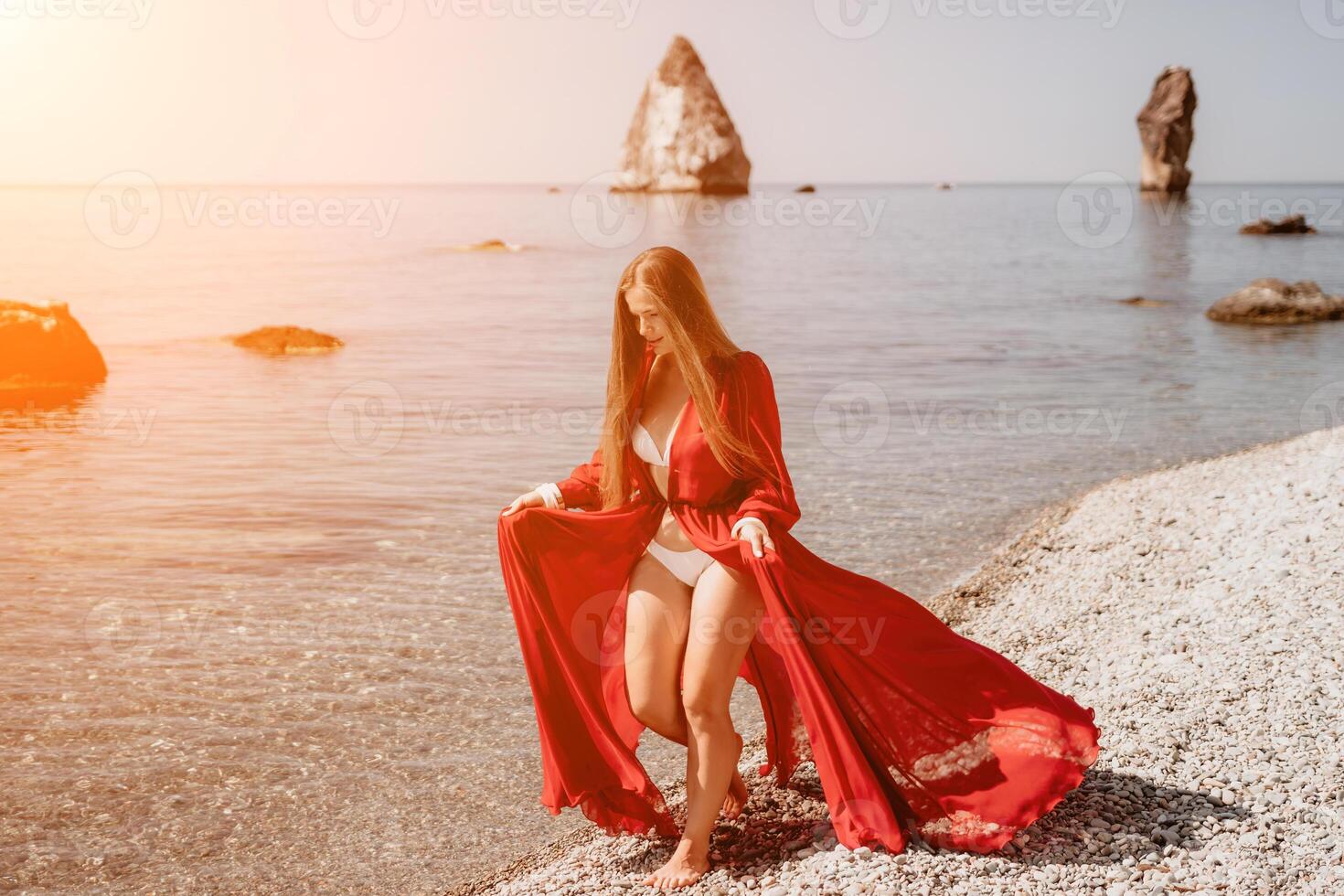 Woman travel sea. Happy tourist in red dress enjoy taking picture outdoors for memories. Woman traveler posing on the rock at sea bay surrounded by volcanic mountains, sharing travel adventure journey photo