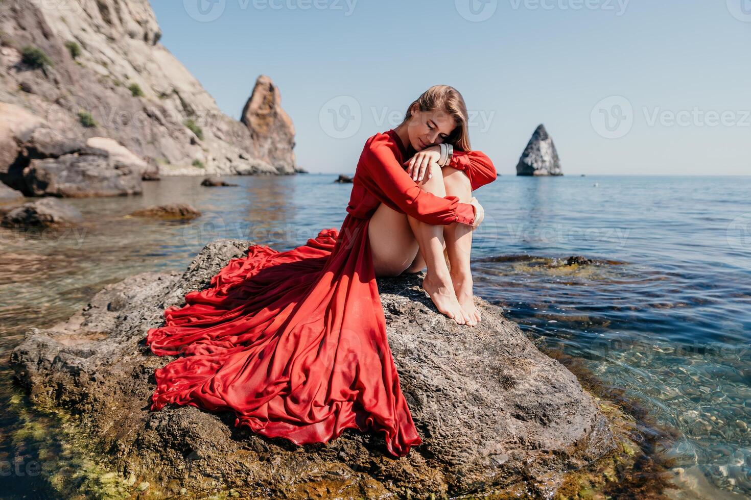 Woman travel sea. Happy tourist in red dress enjoy taking picture outdoors for memories. Woman traveler posing on the rock at sea bay surrounded by volcanic mountains, sharing travel adventure journey photo