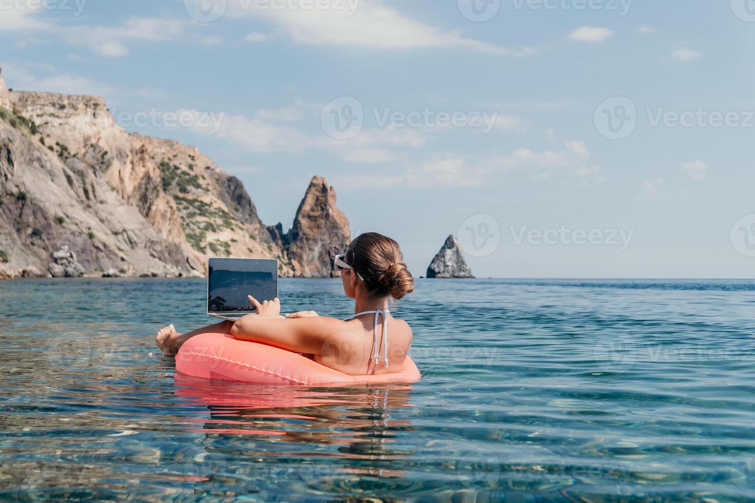 Woman freelancer works on laptop swimming in sea on pink inflatable ring. Happy tourist in sunglasses floating on inflatable donut and working on laptop computer in calm ocean. Remote working anywhere photo