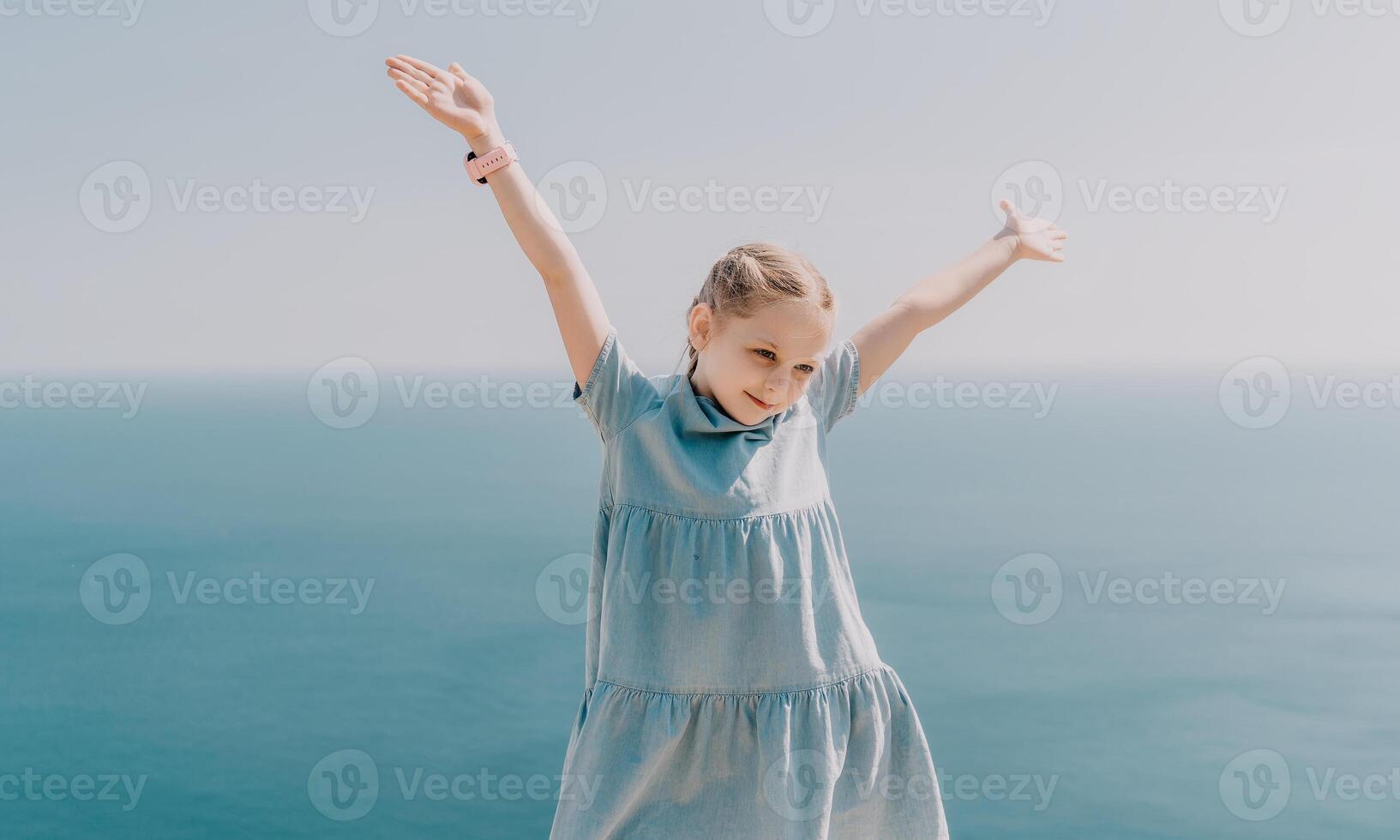 A young girl is standing on a beach, wearing a blue dress and holding her arms up in the air. Concept of joy and freedom, as the girl appears to be enjoying her time by the ocean. photo