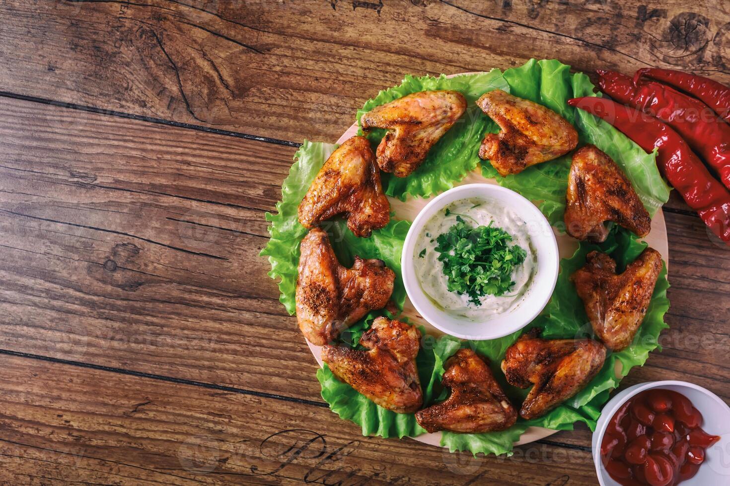 delicious grilled chicken wings with garlic and tomato sauce with lettuce on a round board on wooden rustic background top view, witi copyspace photo