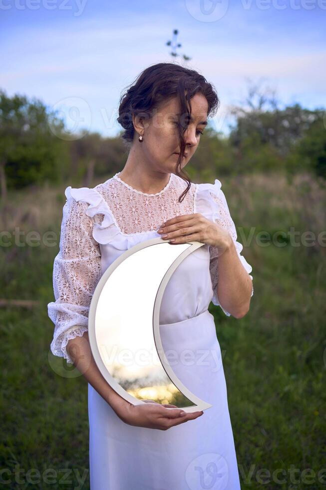 a beautiful woman in a white vintage dress with a train holds a mirror in the shape of the moon photo