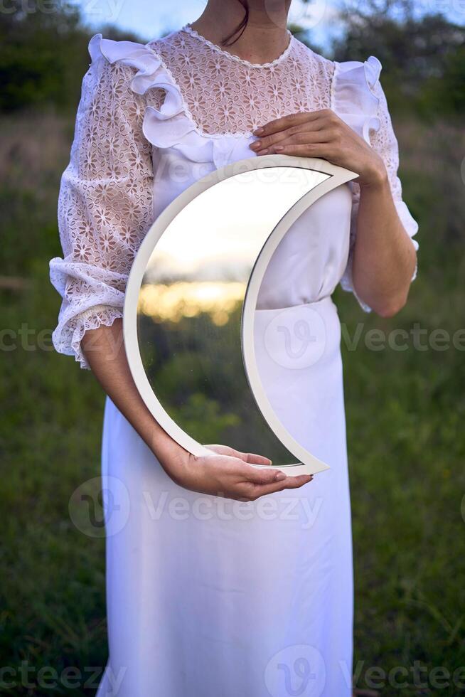 a beautiful woman in a white vintage dress with a train holds a mirror in the shape of the moon photo