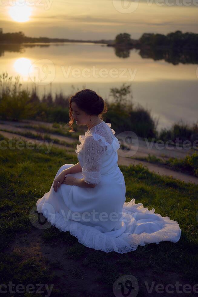 a woman in a white vintage dress on the shore of a lake at sunset photo