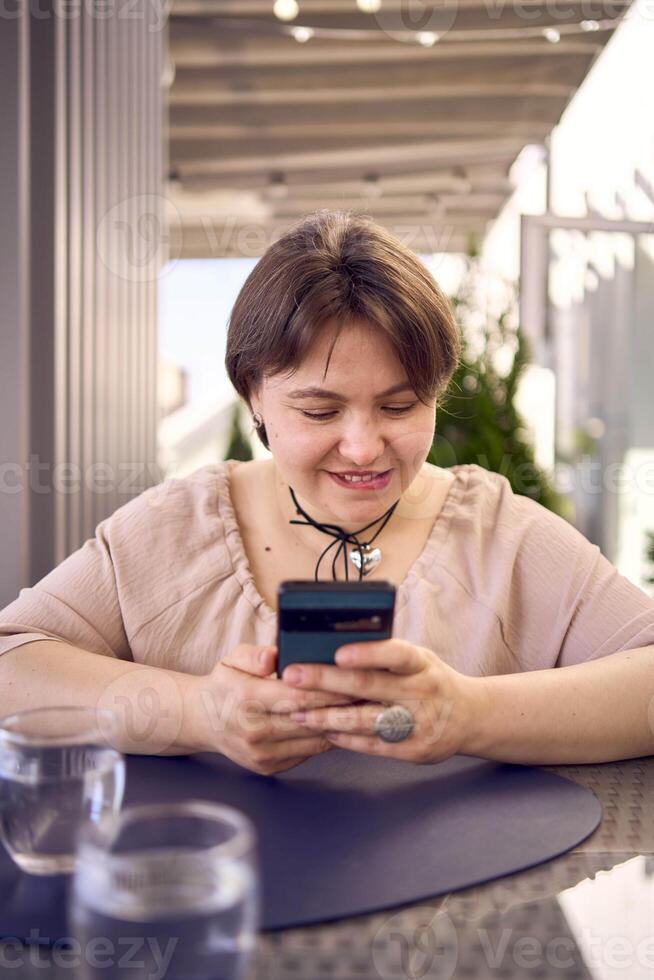 plus size woman in peach fuzz dress chooses a dish in a restaurant via QR code in smartphone, water with lemon and glasses in front of her photo