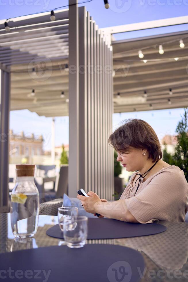 plus size woman in peach fuzz dress chooses a dish in a restaurant via QR code in smartphone, water with lemon and glasses in front of her photo