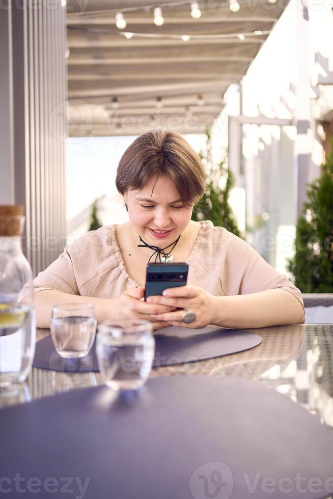 plus size woman in peach fuzz dress chooses a dish in a restaurant via QR code in smartphone, water with lemon and glasses in front of her photo