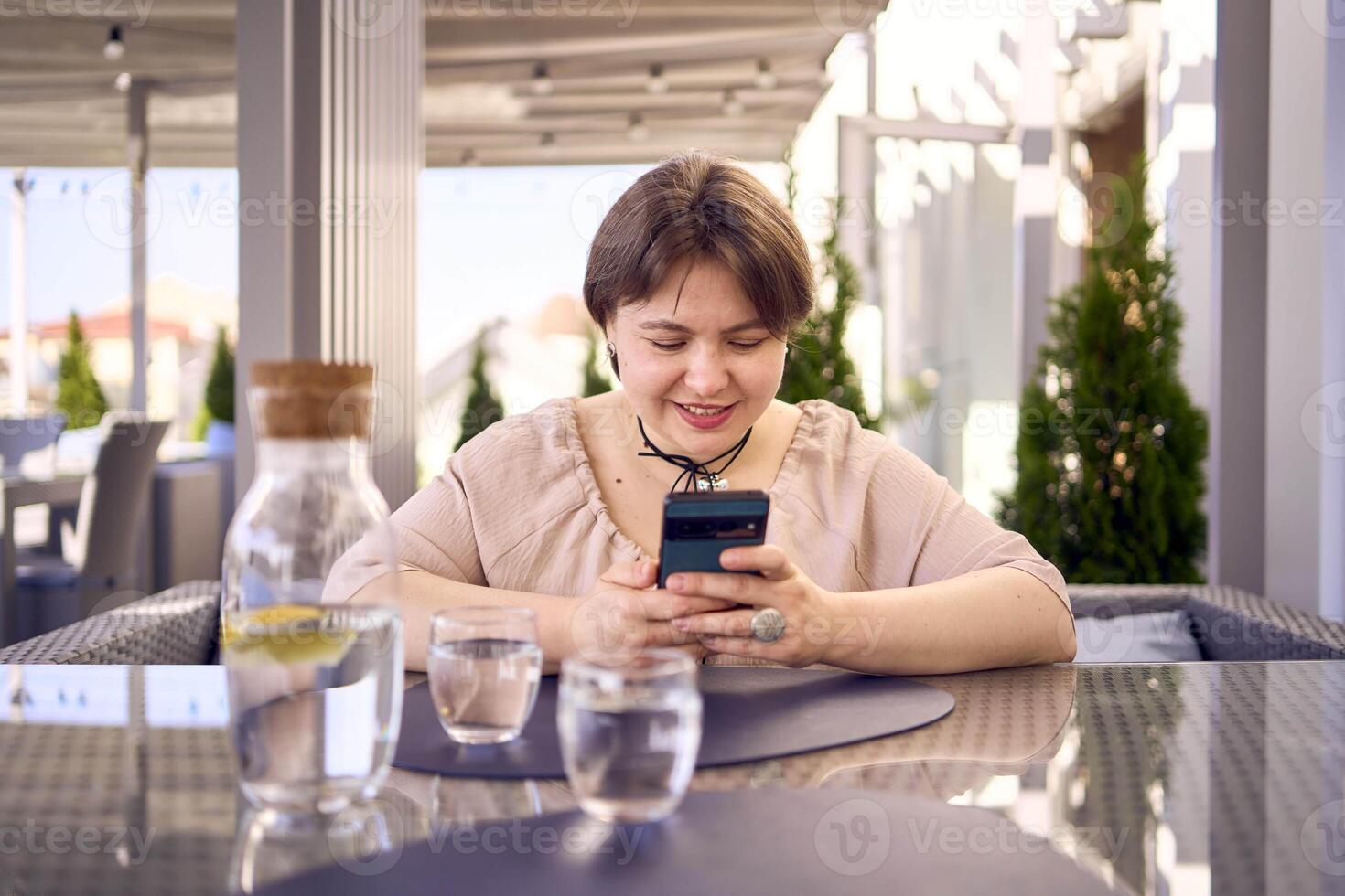 plus size woman in peach fuzz dress chooses a dish in a restaurant via QR code in smartphone, water with lemon and glasses in front of her photo