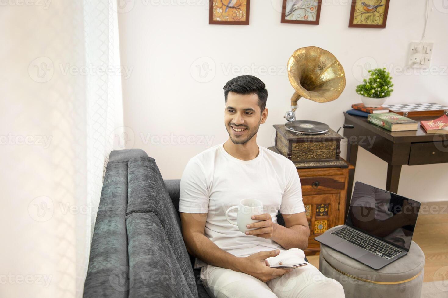 Smiling Hot Indian Man In His Own Thought Holding White Mug Sitting On Sofa photo