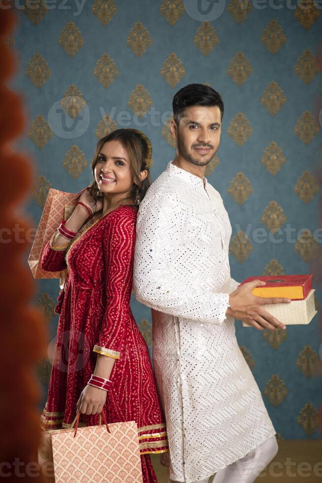 A Stock Photo Of A True-Love Couple Posing For The Camera While Holding Diwali Gifts In Their Hands And Standing Back To Back In Traditional Indian Attire