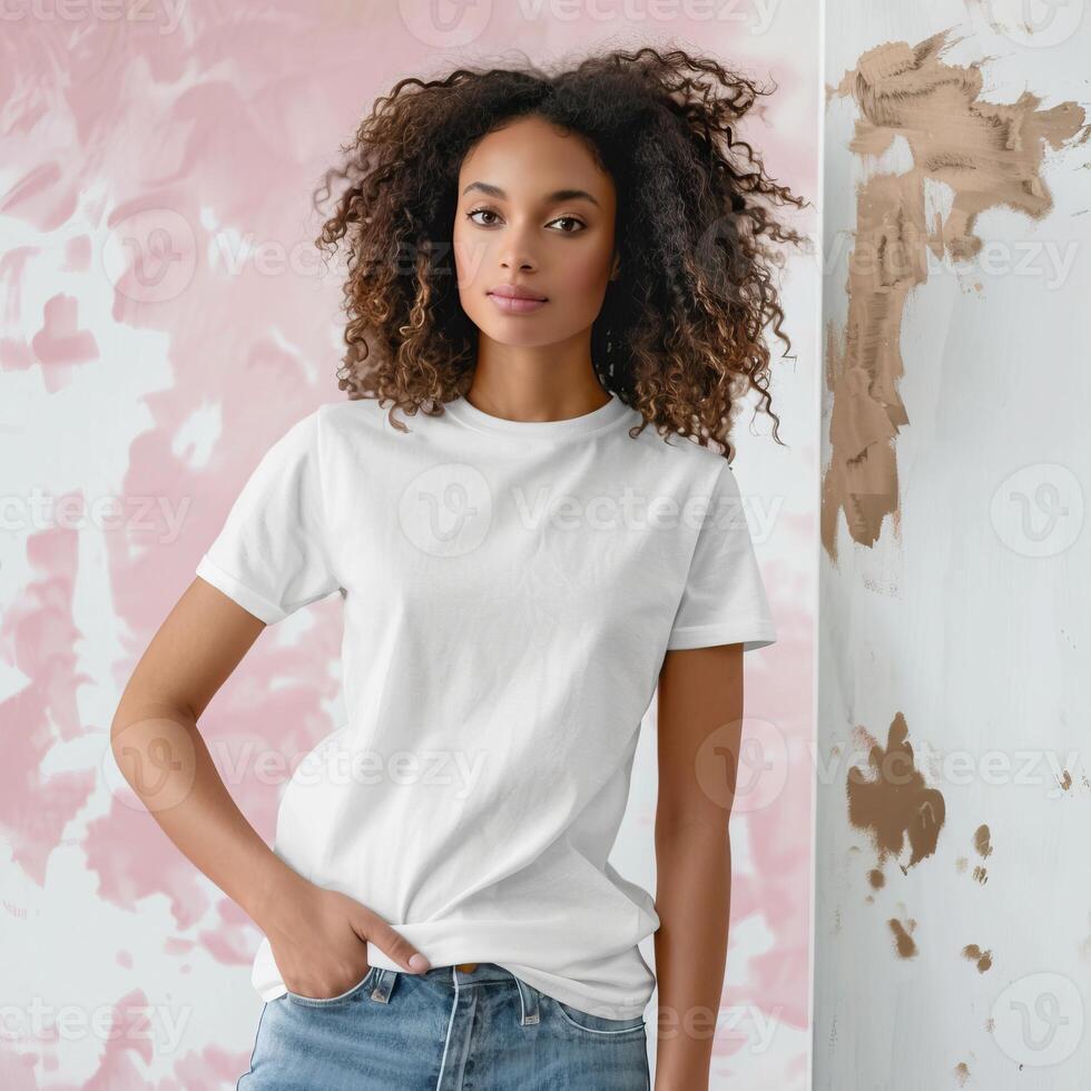 A young woman with long curly hair standing in front of a pink and white background wearing a white t-shirt and blue jeans. photo