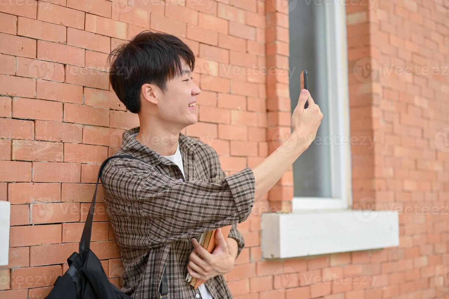 A happy Asian male college student stands against a brick wall of a building using his smartphone. photo