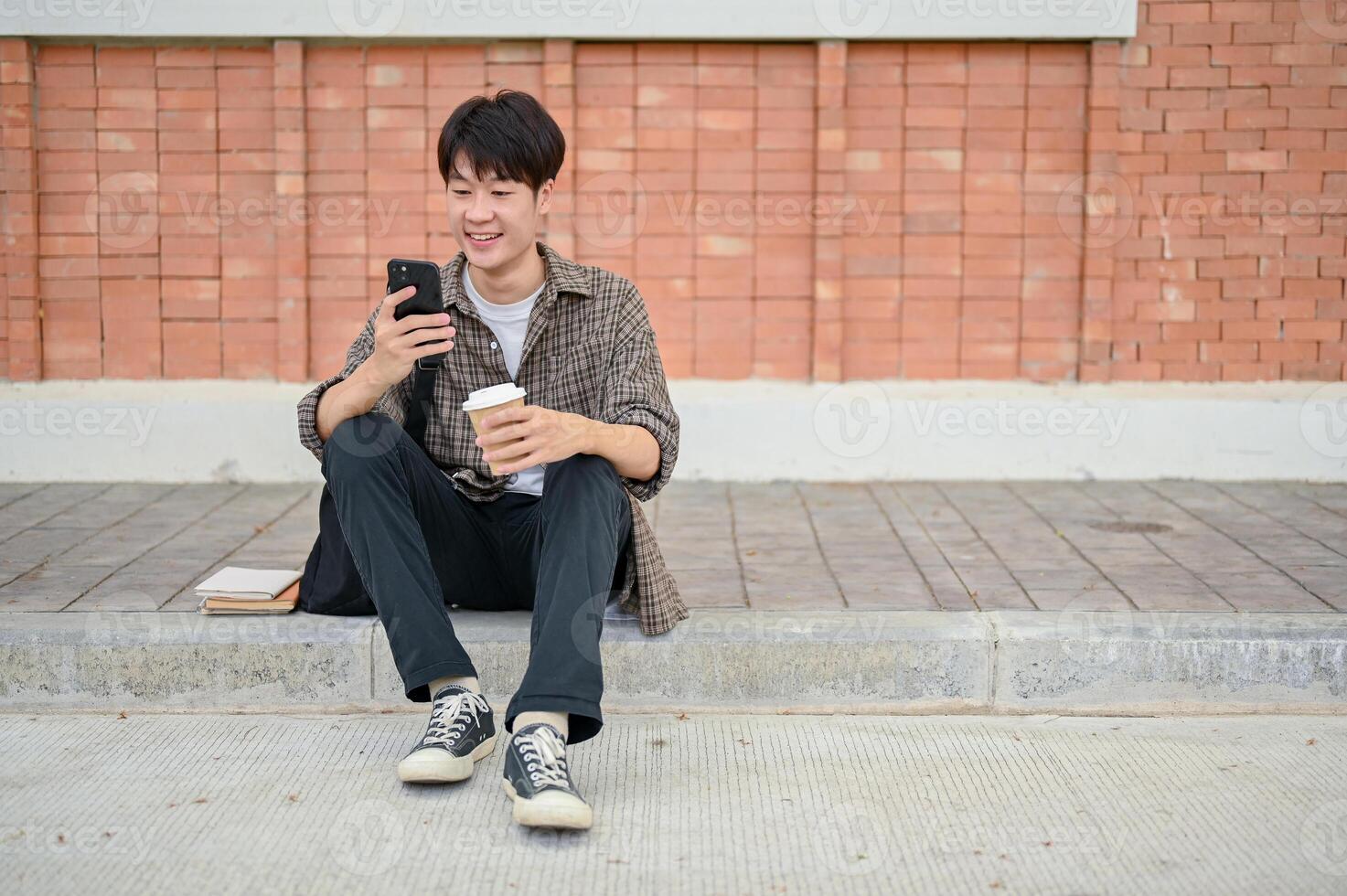 A happy Asian male college student sits on the street in a campus, using his smartphone. photo