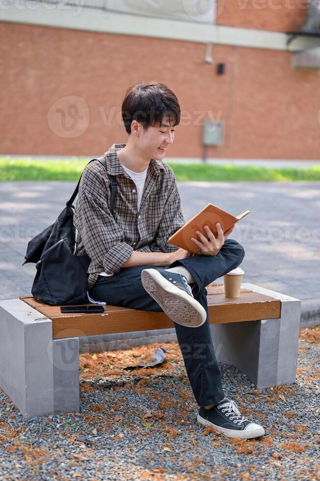 A happy, relaxed Asian male college student sits on a bench in the campus park reading a textbook. photo