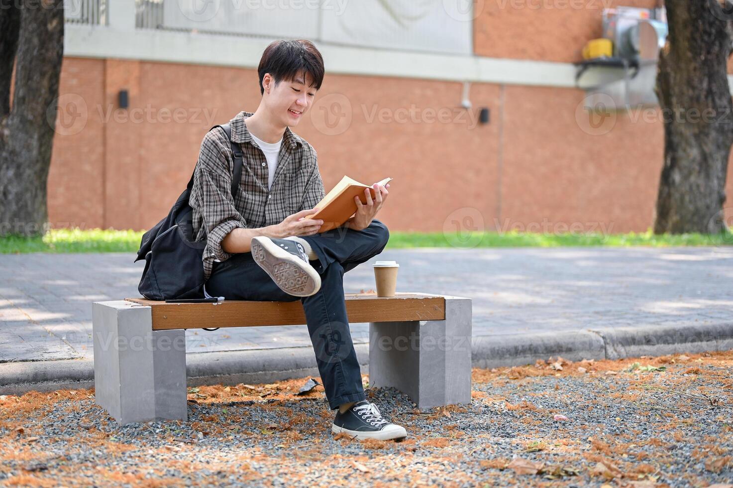 A happy, relaxed Asian male college student sits on a bench in the campus park reading a textbook. photo