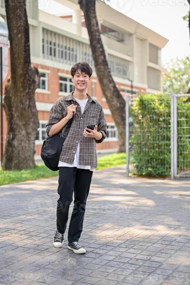 A happy Asian male college student is using his smartphone while walking in the campus park. photo