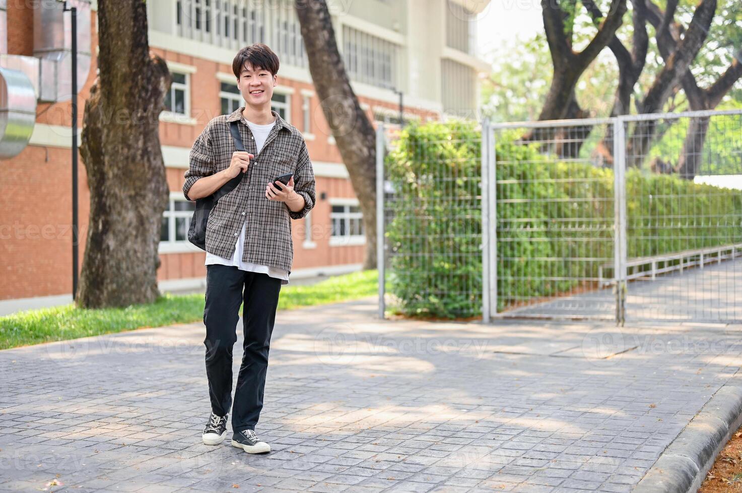 A happy Asian male college student is using his smartphone while walking in the campus park. photo