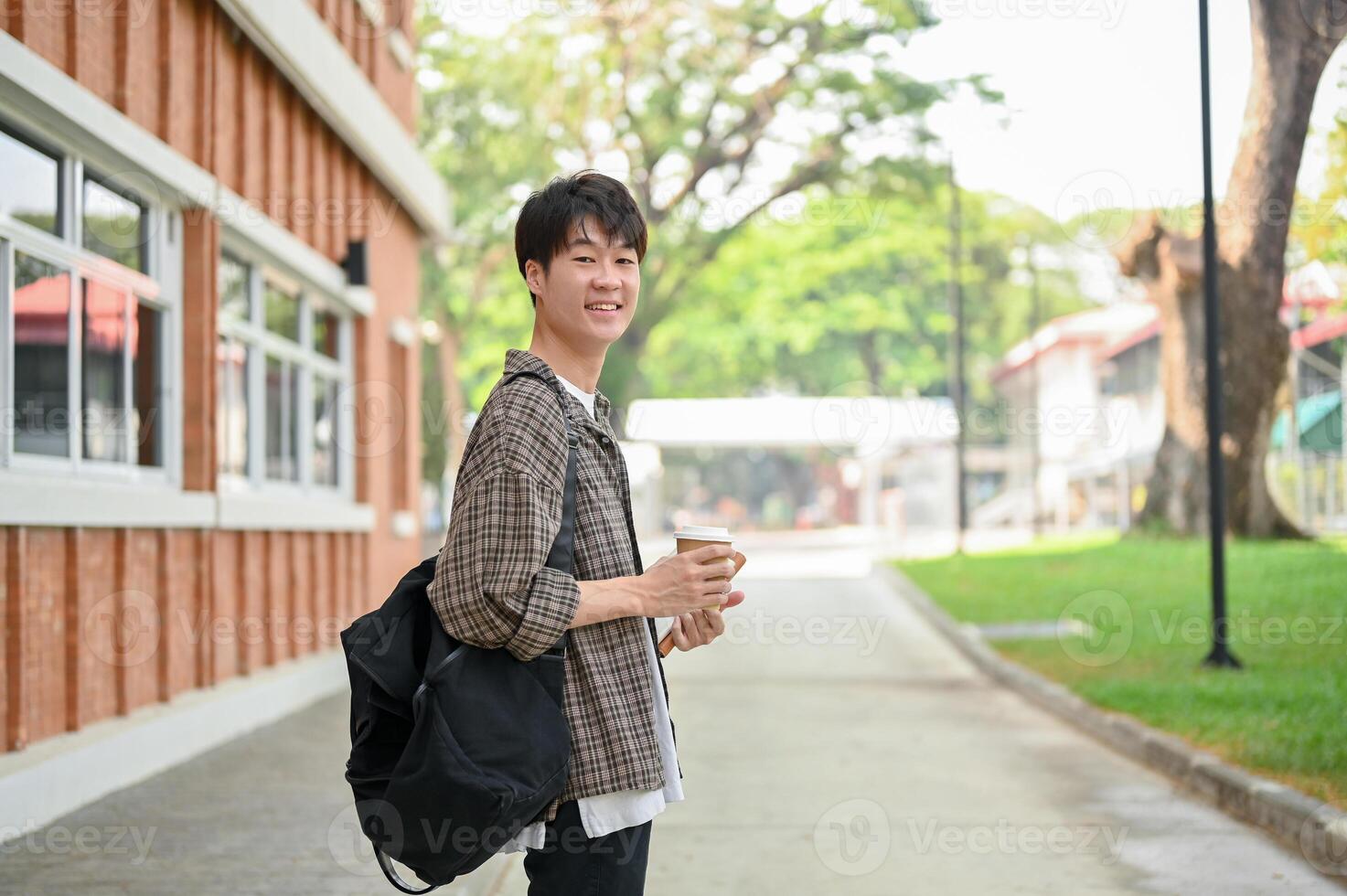 A young Asian man college student with a backpack stands in front of a brick building on campus. photo