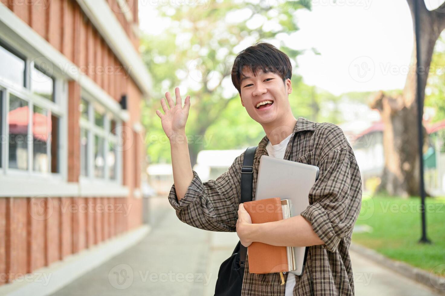 A friendly Asian male student stands outdoors on campus, waving his hand and smiling at the camera. photo