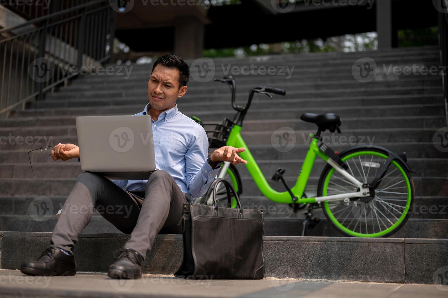 A confused Asian businessman sits on steps with a laptop, showing puzzled expression. photo