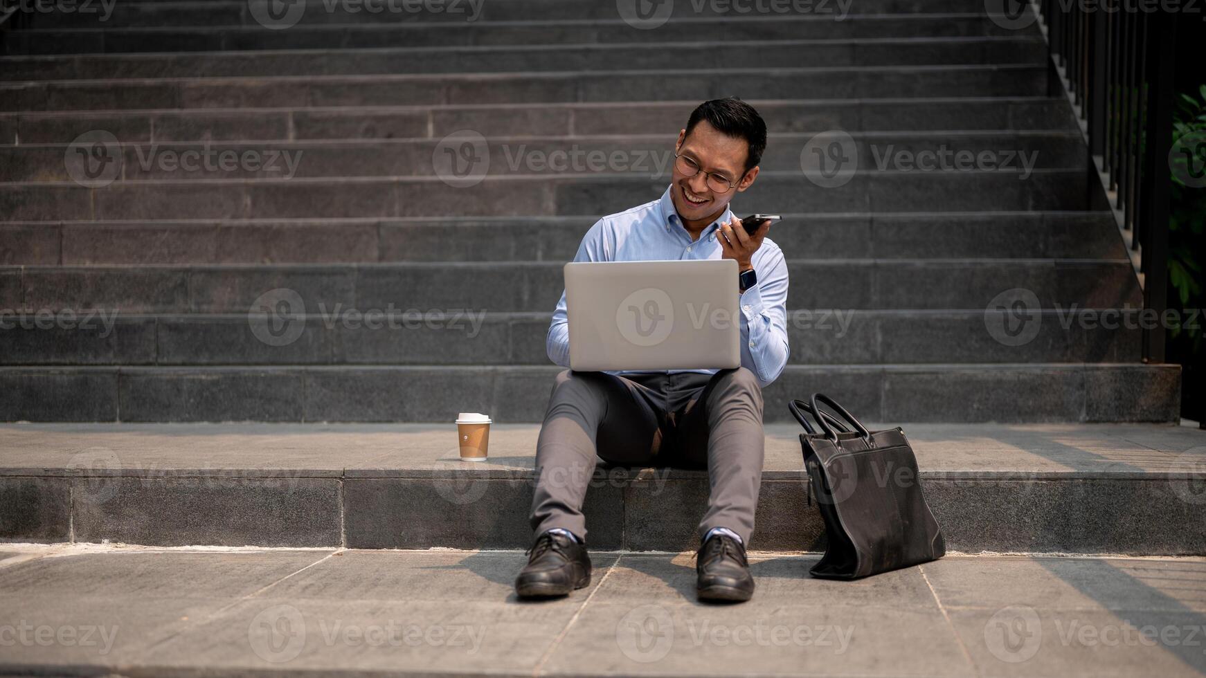 A businessman sits on steps, sending voice message via his smartphone while working on his laptop. photo