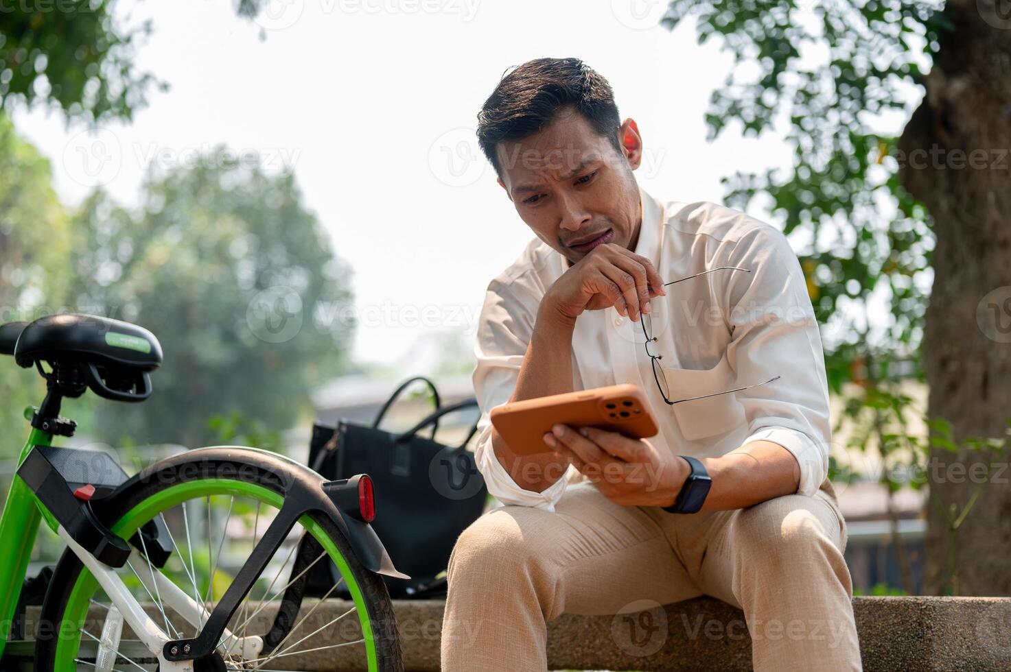 Sad Asian businessman sits outdoors, holding his phone with a concerned expression, deep in thought. photo
